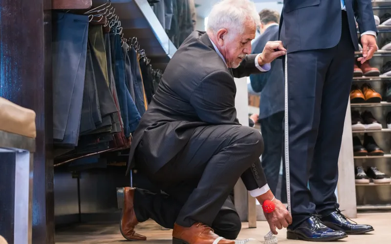 Een man laat zijn pak aanmeten. Je ziet de kleermaker en de benen van de man in pak