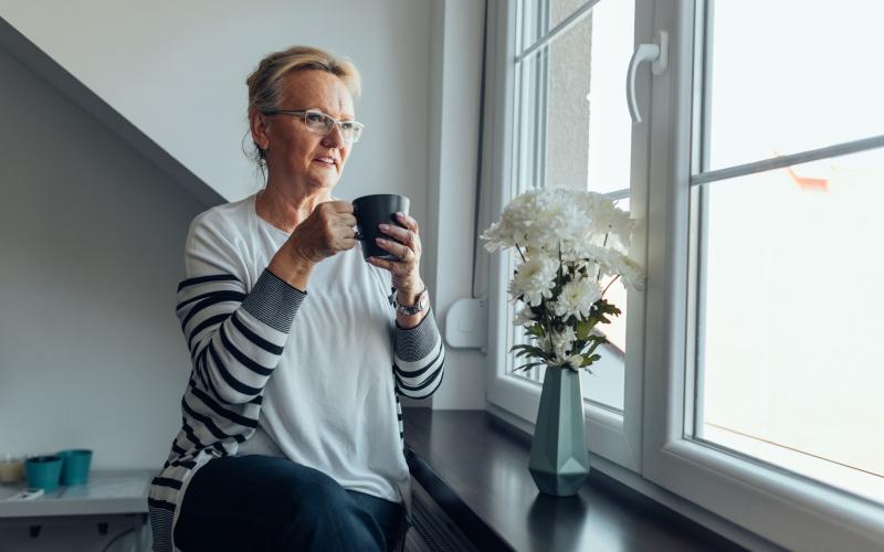 Senior vrouw zit bij het raam en heeft een mok in haar hand