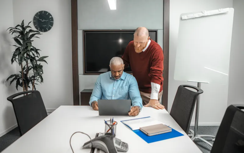 Twee collega's bij elkaar hangen over een laptop. Ze denken na.