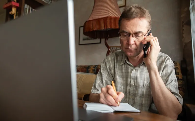 Een man zit aan tafel en is aan het bellen. Ondertussen schrijft hij iets op in een notitieboekje
