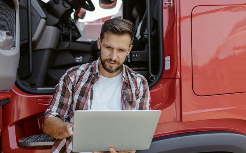 Man staat tegen zijn vrachtwagen aan en heeft een laptop vast Man staat tegen zijn vrachtwagen aan en heeft een laptop vast