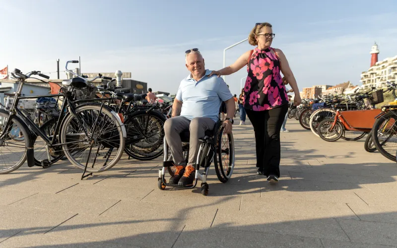Ouder stel loopt langs het strand. Een vrouw heeft haar hand op de schouder van haar partner, die in een rolstoel zit