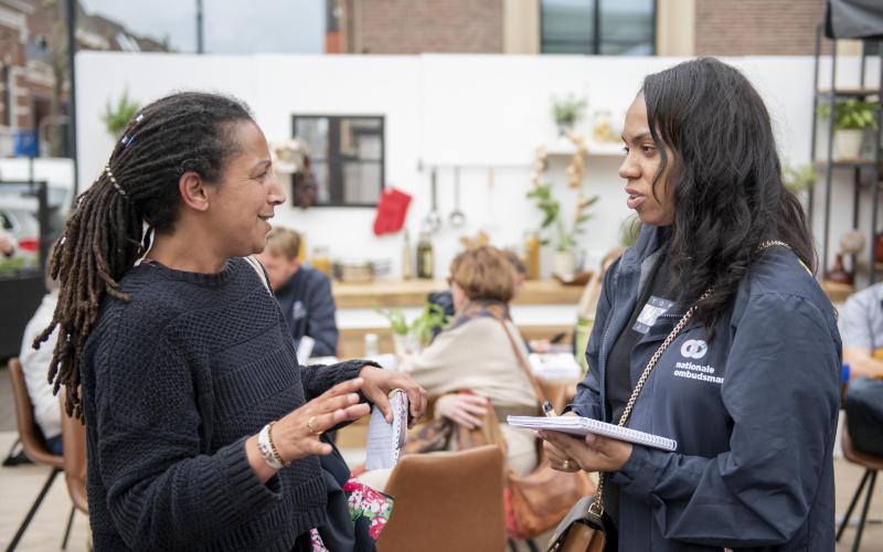 Twee vrouwen staan op een plein voor de pop-upkeuken van de Nationale ombudsman. Ze zijn in gesprek. Twee vrouwen staan op een plein voor de pop-upkeuken van de Nationale ombudsman. Ze zijn in gesprek.