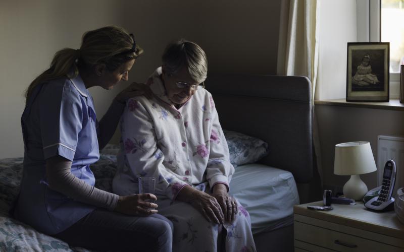 Oudere dame zit op bed zit samen met een verpleegster. De verpleegster slaat een arm om de vrouw heen.