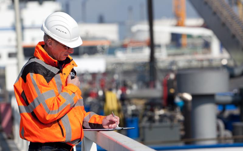 Foto van een man in een oranje veiligheidsjas en met een witte helm op. Hij staat voor een haven. Foto van een man in een oranje veiligheidsjas en met een witte helm op. Hij staat voor een haven.