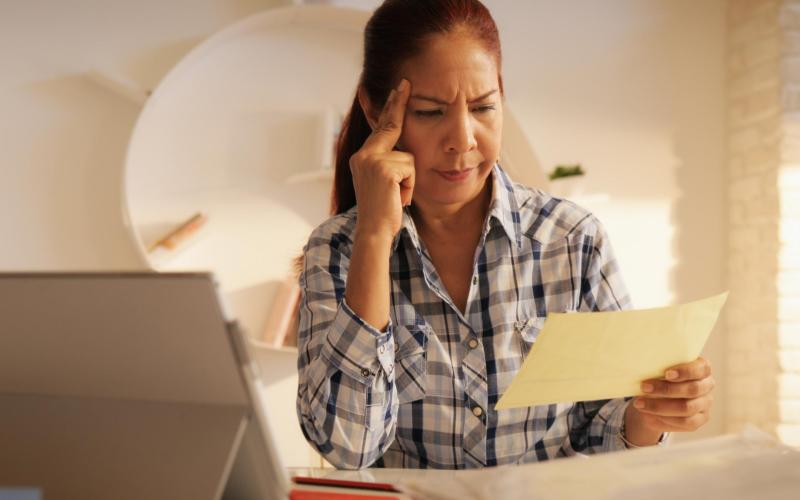 Vrouw zit aan tafel met laptop en kijkt fronsend naar brief in haar hand