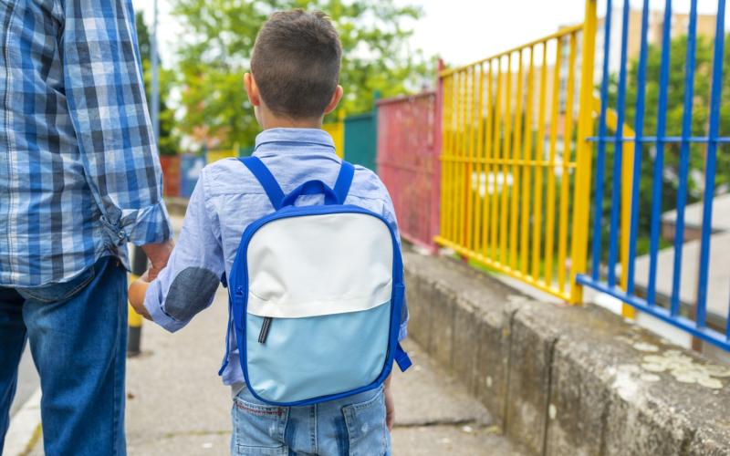 Jongetje met blauw shirt en rugzak loopt bij z'n vader aan de hand