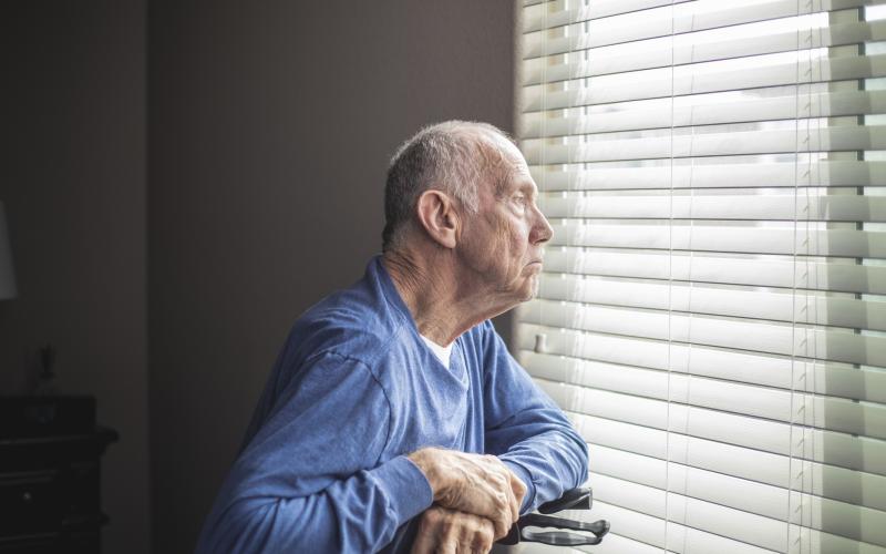 Een oudere man leunt op zijn rollator en kijkt uit het raam