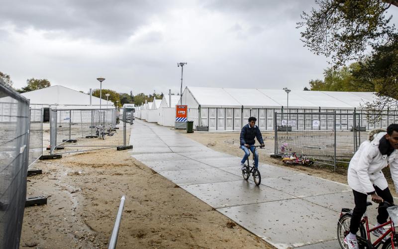 Twee mannen fietsen over het AZC terrein van Heumensoord. Tenten op de achtergrond