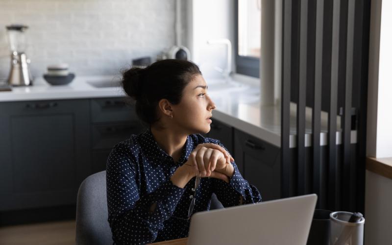 Vrouw met donker haar, zit in woonkamer en kijkt peinzend naar buiten door het raam Vrouw met donker haar, zit in woonkamer en kijkt peinzend naar buiten door het raam