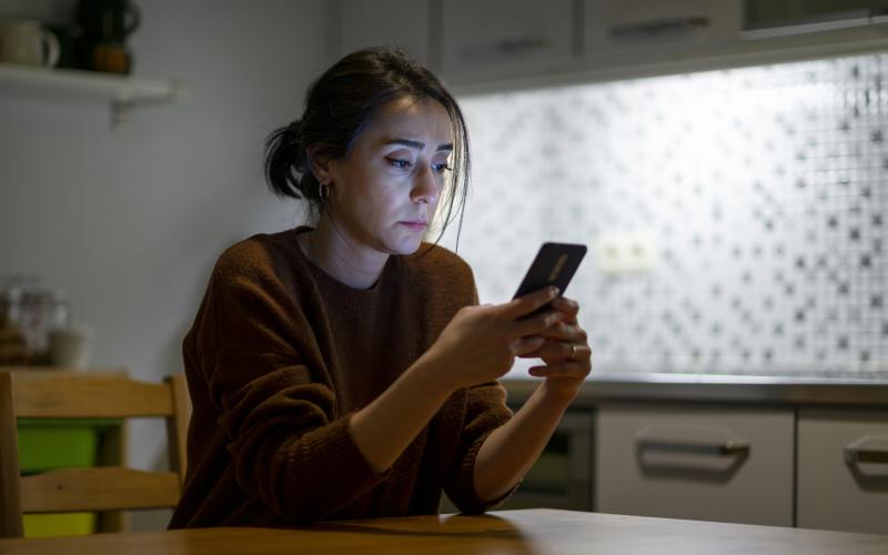 Vrouw zit aan tafel in haar keuken en kijkt peinzend op haar smartphone
