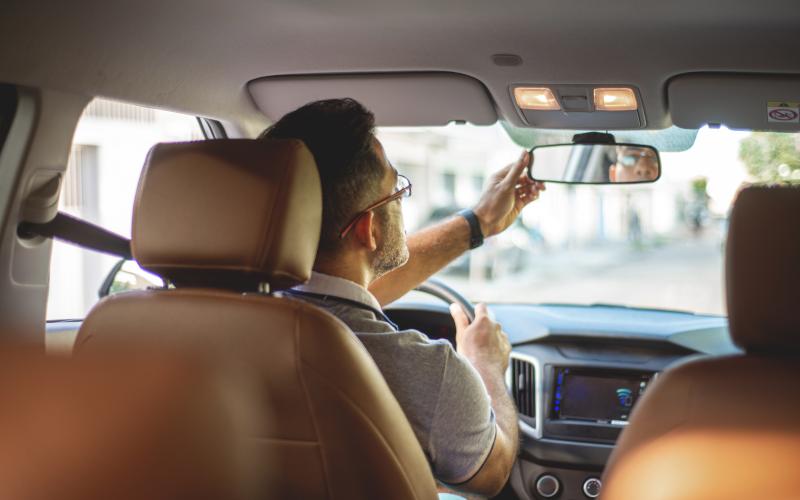 Man in auto op bestuurdersstoel van achteren gefotografeerd. Hij houdt zijn achteruitkijkspiegel vast en kijkt daar in.