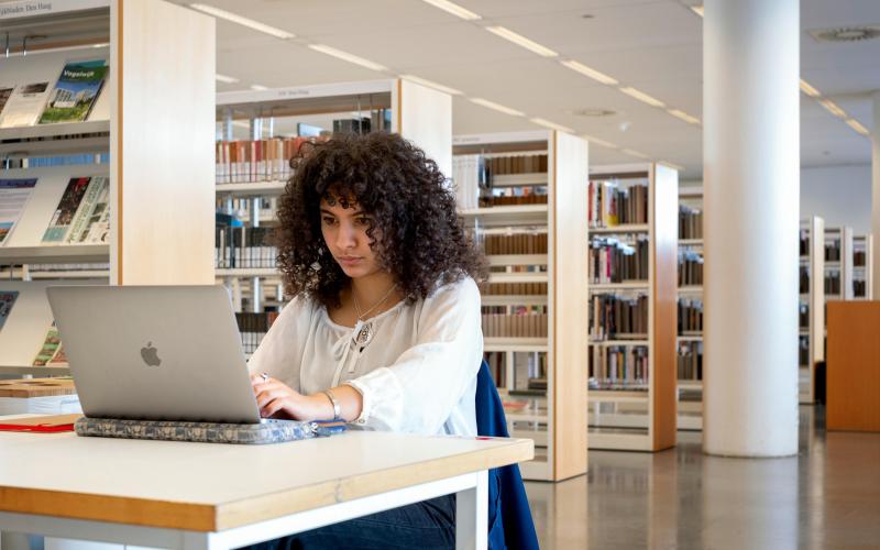 Jonge vrouw zit aan een tafeltje in een bibliotheek met haar laptop voor zich opengeklapt.