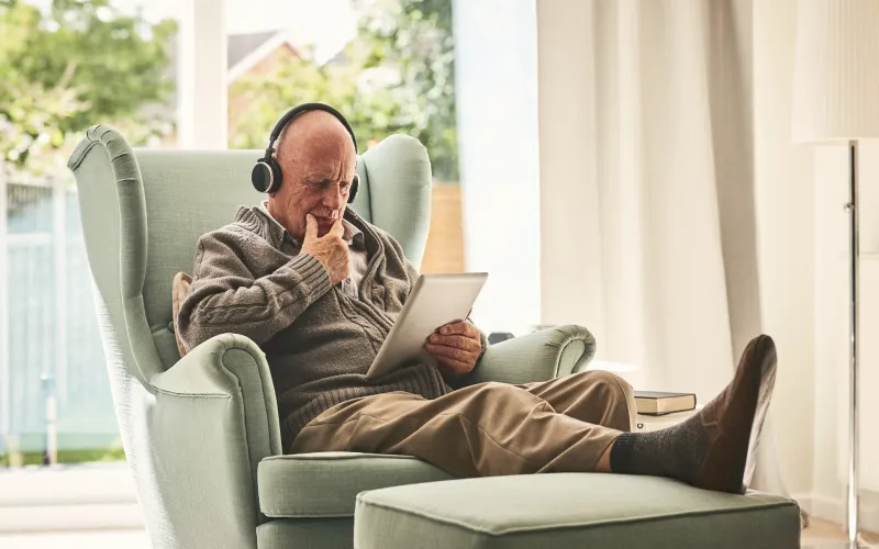 Oudere man zit op stoel met koptelefoon op zijn hoofd en tablet in zijn hand.