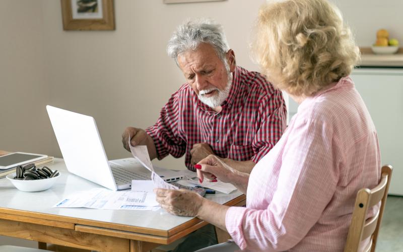Ouder echtpaar zit aan tafel met een laptop en papieren voor zich. De man kijkt zorgelijk.