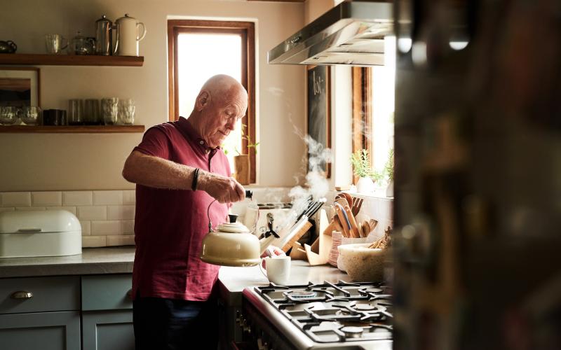 Oudere man staat in de keuken en houdt een theepot vast. Hij schenkt het in een kopje.