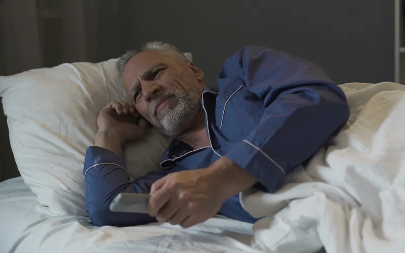 Een oudere man ligt wakker in bed. Hij heeft zijn smartphone vast.