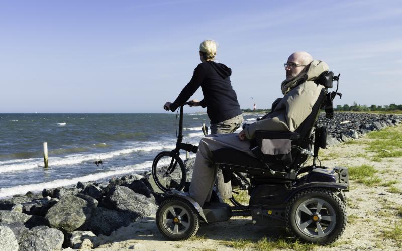 Een senior man zit in een scootmobiel. Naast hem staat een vrouw met een fiets. Ze staan op de rotsen dicht bij zee.