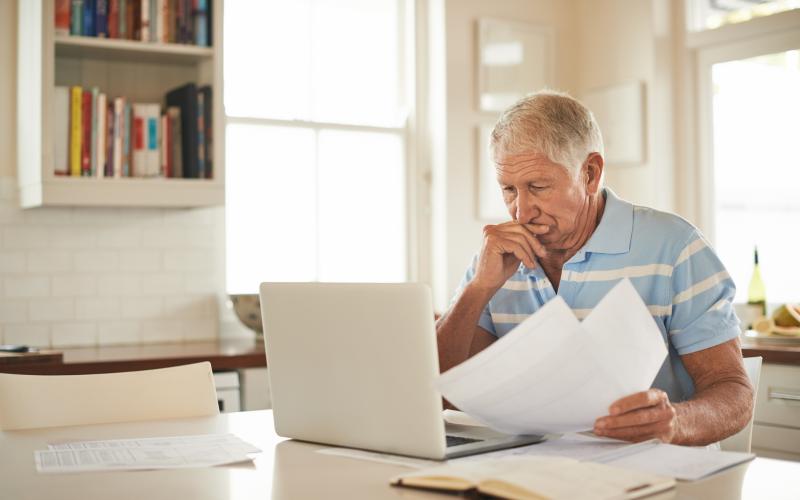 Een oudere man zit bezorgd achter zijn computer met papieren in zijn hand