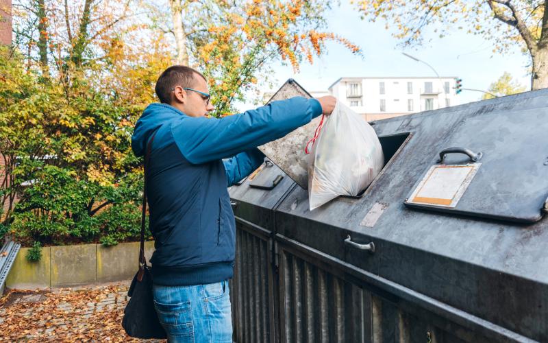 Een man gooit een zak vuilnis in een afvalcontainer. Hij draagt een jas, schoudertas en heeft een bril op.