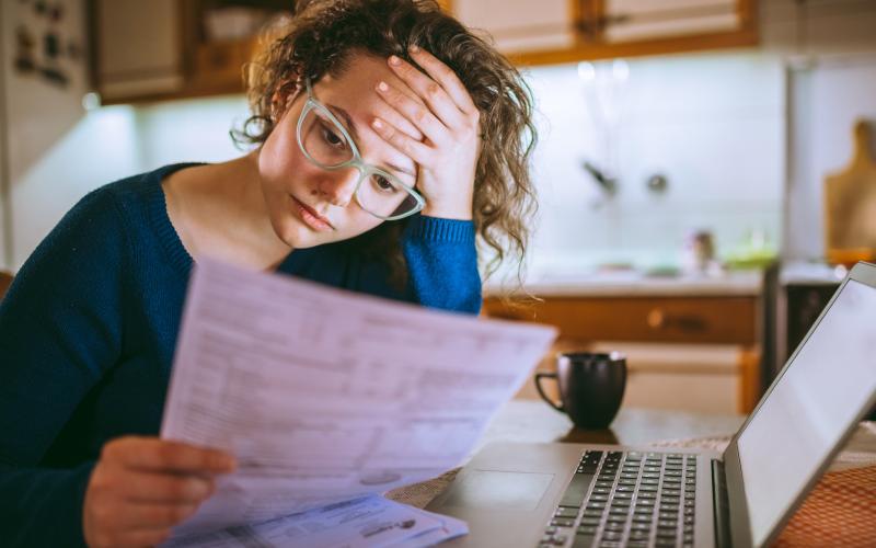 Een vrouw zit met papieren in haar hand aan tafel. Voor haar staat een laptop. Ze kijkt zorgelijk. Ze draag een bril en heeft een blauwe trui aan.