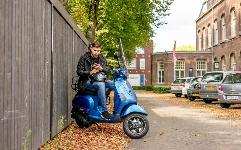 Jongere zit op zijn scooter. Hij staat stil en kijkt op zijn smartphone.