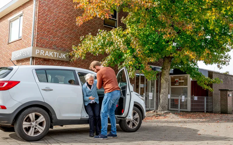 Mantelzorger helpt oudere vrouw uit de auto. Ze staan geparkeerd voor de huisartsenpraktijk.