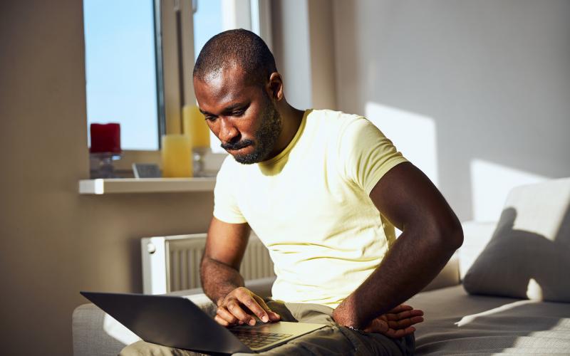 Een jongvolwassen man met geel t-shirt zit op zijn bank en heeft een laptop op zijn schoot. Hij kijkt bedenkelijk.