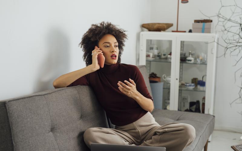 Een volwassen vrouw zit in kleermakerszit op de bank. Ze is aan het bellen.