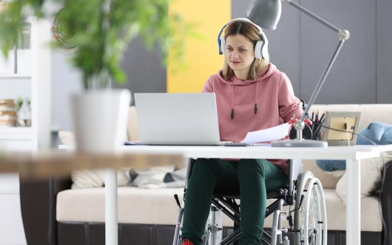 Een jongvolwassen vrouw in een rolstoel zit thuis achter haar laptop te werken of studeren. Ze draagt een roze trui en heeft een koptelefoon op.