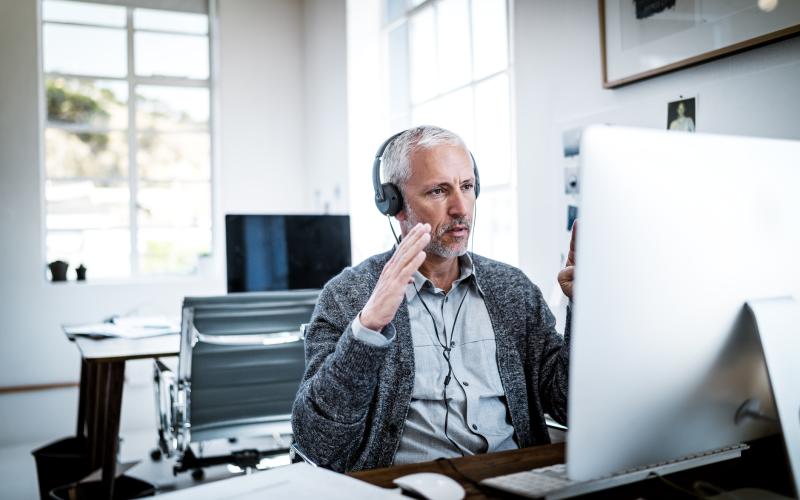 Een man met grijs haar zit te werken achter zijn computer. Hij draagt een koptelefoon.