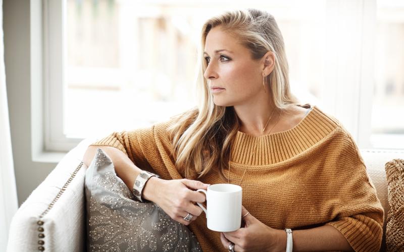 Vrouw van middelbare leeftijd met lang blond haar zit op de bank met een kop koffie in haar hand. Ze kijkt voor zich uit.