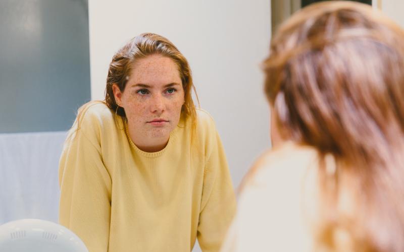 Een jongvolwassen vrouw staat voor de spiegel en kijkt zichzelf aan. Ze heeft lang blond haar en draagt een gele trui.