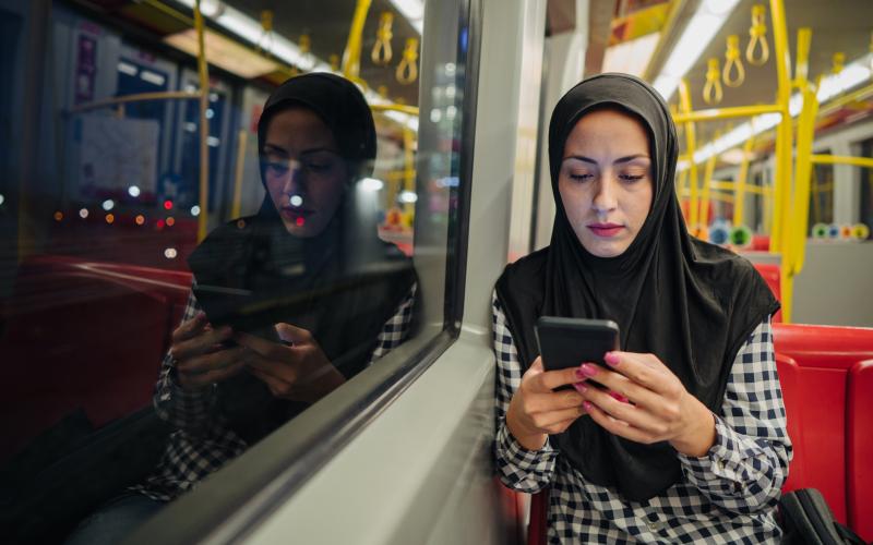 Een vrouw van middelbare leeftijd met hoofddoek en geblokte blouse zit met haar telefoon in haar hand in het openbaar vervoer.