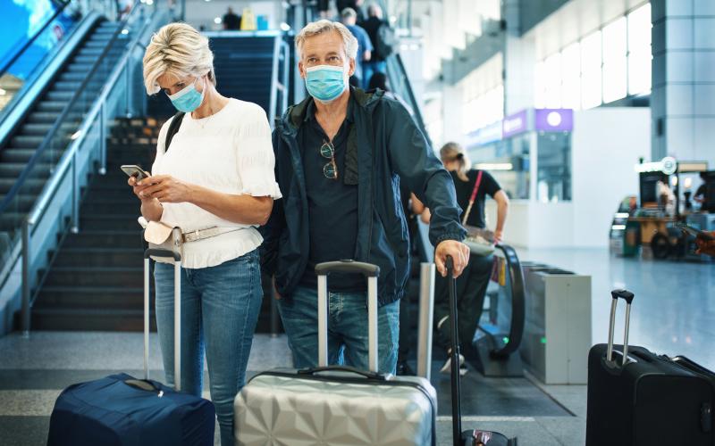 Ouder echtpaar staat op het vliegveld met koffers voor zich. Ze dragen allebei een mondkapje.