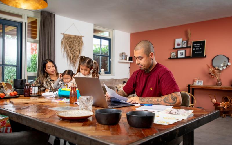 Jong gezin van vader, moeder en twee jonge dochters regelt thuis zaken achter laptop. Het is een kleurrijke omgeving.