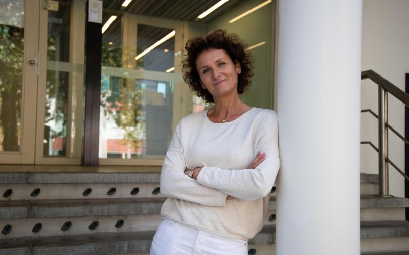 Vrouw met donkere krullen (Emily Govers, Nationale ombudsman) draagt een witte broek en shirt. Ze leunt tegen een wand Vrouw met donkere krullen (Emily Govers, Nationale ombudsman) draagt een witte broek en shirt. Ze leunt tegen een wand