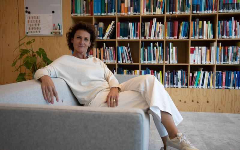 Vrouw met donkere krullen zit op de bank. Achter haar staat een boekenkast Vrouw met donkere krullen zit op de bank. Achter haar staat een boekenkast