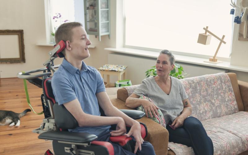 Een jongen in een elektrische rolstoel in gesprek met een vrouw op de bank ernaast