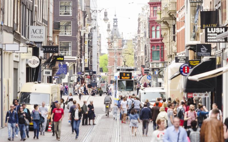 Een foto van een straat in Amsterdam. Je ziet mensen wandelen, tramrails, en aan beide kanten winkels.
