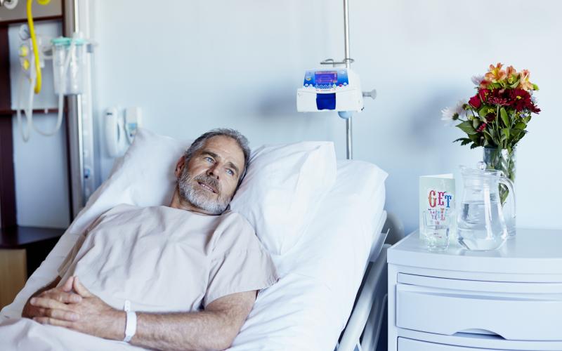 Man ligt in ziekenhuisbed. Hij heeft een baard en draagt een licht gekleurd shirt. Naast het bed staan bloemen.