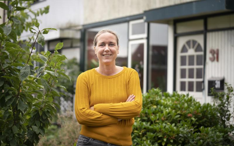 Vrouw met gele trui staat glimlachend voor haar huis