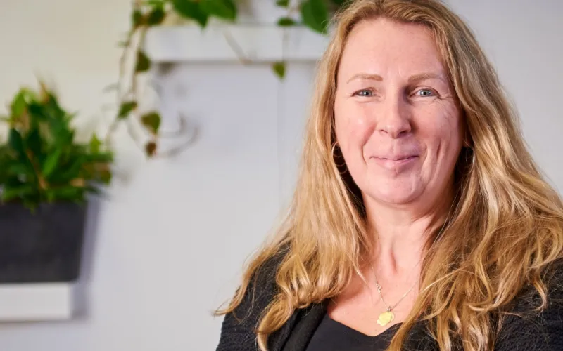 Glimlachende vrouw (Sociaal Werker Marloes Olde Hampsink) met lang blond haar, zwart shirt. Op de achtergrond staan planten