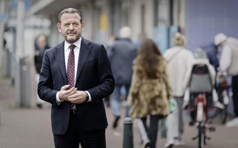 Man met baard (Reinier van Zutphen, Nationale ombudsman), draagt een pak, handen gevouwen, staat op straat met op de achtergrond winkelend publiek 