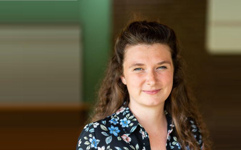 Vrouw (Sophie Albers) heeft lang donker haar en draagt een zwarte blouse met bloemen