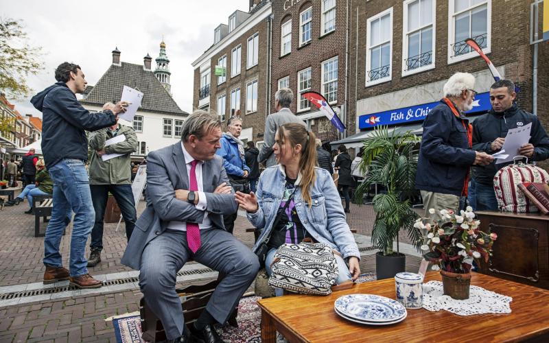 De Nationale ombudsman op bezoek in Zeeland. Hij is in gesprek met een vrouw met donkerbruin lang haar. Ze bevinden zich op een plein.