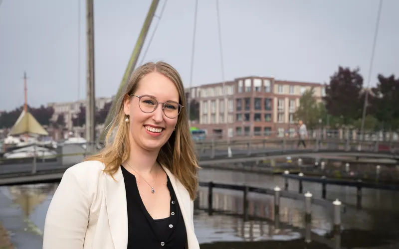 Vrouw (Michelle Koelewijn) heeft blond haar en draagt een bril. Ze staat buiten bij het water (haven)