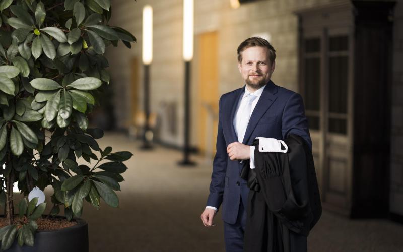 Man (Herman van Harten) heeft baard en draagt donker pak. Hij staat in een gang