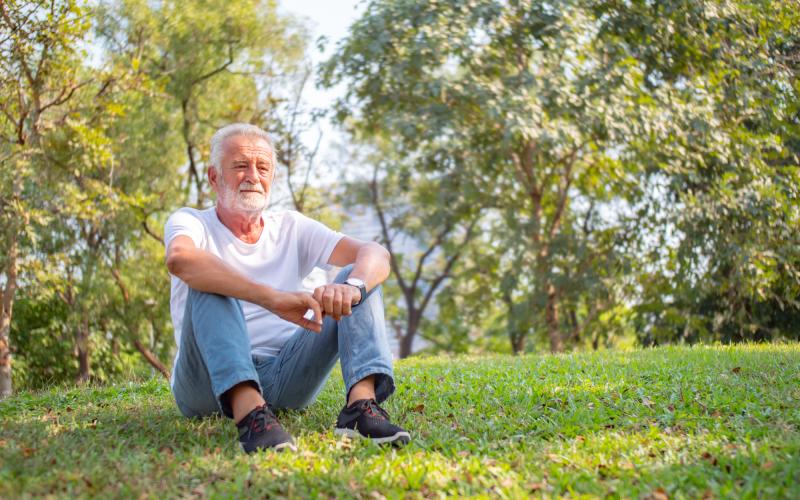 Senior man zit op grasveld. Hij heeft grijs haar en een grijze baard. Hij draagt een wit shirt en spijkerbroek