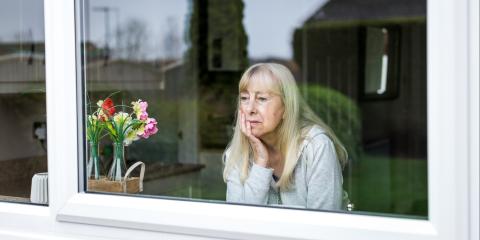 Oudere vrouw zit achter het raam en kijkt naar buiten. Ze kijkt zorgelijk en steunt haar hoofd op haar hand.